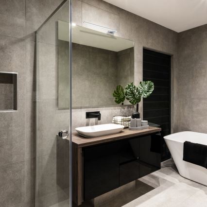 Moderná kúpeľňa s umývadlom na tmavo-hnedej drevenej skrínke. Umývadlo je umiestnené pod zrkadlom s LED osvetlením. V pozadí je sprchovací kút a voľne stojaca vaňa.