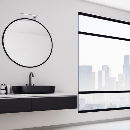 Okrúhle zrkadlo visí na stene kúpeľne. Na umývadle s čiernou umývadlovou misou tečie voda z kohútika. Za umývadlom sa nachádza veľké okno s výhľadom na mestskú panorámu.