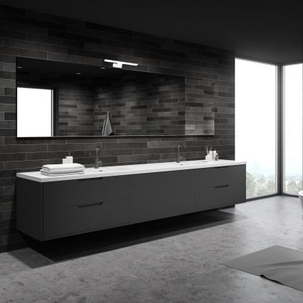 Dvojité umývadlo s dvoma batériami je umiestnené na sivej skrinkovej skrinke v kúpeľni. Steny sú obložené tmavými dlaždicami, nad umývadlom je veľké zrkadlo s osvetlením. Podlaha je betónová, v pozadí je veľké okno s výhľadom do prírody.