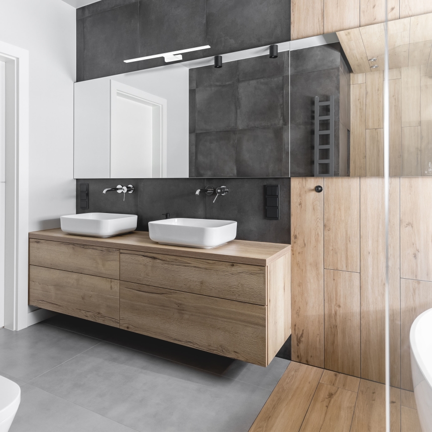 Dve umývadlá na drevenom nábytku sú umiestnené pod zrkadlom v modernej kúpeľni s tmavosivými a svetlohnedými obkladmi.