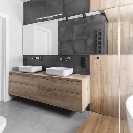 Dve umývadlá sú umiestnené na drevenom nábytku v kúpeľni. Nábytok je súčasťou kúpeľňového zariadenia s tmavosivými dlaždicami a svetlými drevenými panelmi.