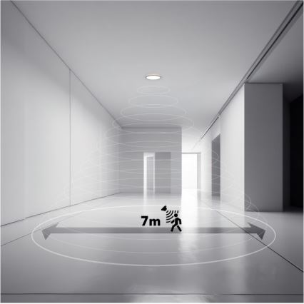 Svietidlo s pohybovým senzorom detekuje pohyb v priestore dlhej chodby. Dosah detekcie je 7 metrov. Chodba je svetlá, s bielymi stenami a dverami.