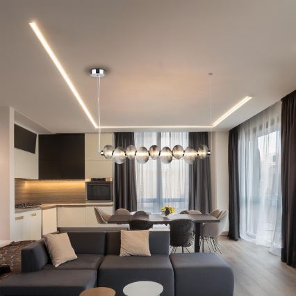 Dlhá prívesná lampa s guľatými sklenenými prvkami visí nad jedálenským stolom v otvorenej kuchyni a obývacej izbe. Lampa je umiestnená medzi kuchyňou a obývačkou s moderným nábytkom.