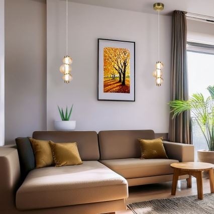 Dve závesné lampy s tromi sklenenými guľami visia v obývacej izbe. Svetlohnedá rohová sedačka s vankúšmi je umiestnená pred nimi. V pozadí je obraz stromov a izbová rastlina.