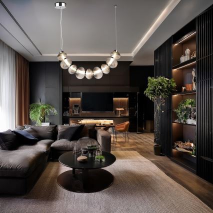 Moderné stropné svietidlo s krivkou z priehľadných sklenených guľôčok visí v tmavej obývacej izbe nad hnedou sedacou súpravou a konferenčným stolíkom. Izba obsahuje aj nábytok z tmavého dreva a rastliny.