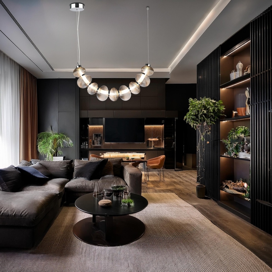 Moderné stropné svietidlo s krivkou z priehľadných sklenených guľôčok visí v tmavej obývacej izbe nad hnedou sedacou súpravou a konferenčným stolíkom. Izba obsahuje aj nábytok z tmavého dreva a rastliny.
