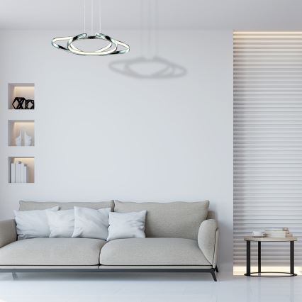 Biela sedačka s tmavými vankúšmi stojí pred stenou so štruktúrovaným povrchom. Nad sedačkou visí moderné stropné svietidlo s troma prepletenými kruhmi.