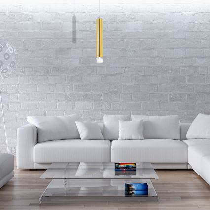 Zlatá závesná lampa visí nad bielou poličkou. Na poličke sú knihy, vázy, sviečky a dekorácie. Polička je umiestnená v modernom interiéri s krbom a policami s ozdobnými fľašami.