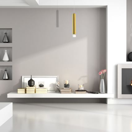 Zlatý valcový závesný svietidlo visí na tenkom kábli. Spodná časť svietidla je ukončená priesvitným valčekom. Biele pozadie.