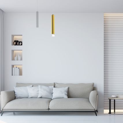 Zlaté valcovité svietidlo visí v obývacej izbe. Biela sedačka s vankúšmi stojí pred stenou z bielych tehál. Na priehľadnom konferenčnom stolíku ležia knihy.