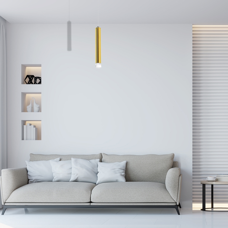 Zlaté valcovité svietidlo visí v obývacej izbe. Biela sedačka s vankúšmi stojí pred stenou z bielych tehál. Na priehľadnom konferenčnom stolíku ležia knihy.