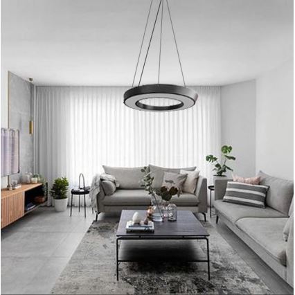 Kruhové stropné svietidlo visí v obývacej izbe. Svietidlo je čierne s vnútorným bielym kruhom. Izba obsahuje sivú sedačku, konferenčný stolík a koberec.