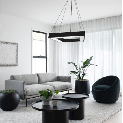 Štvorcové závesné svietidlo svieti v obývacej izbe. Izba obsahuje sivú sedačku, dva nízke stolíky, kreslo a rastliny v kvetináčoch.