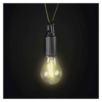 Žiarovka svieti. Je zavesená na šnúrke na tmavofialovom pozadí. Žiarovka má čierny kovový kryt.