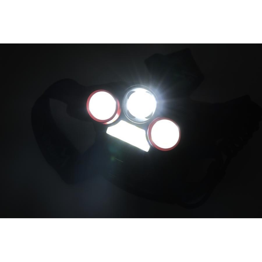 Čelová lampa svieti. Má tri kruhové svetlá a jedno obdĺžnikové. Je umiestnená na tmavom pozadí.