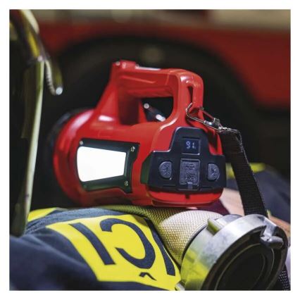 Červená prenosná lampa s displejom ukazuje 96%. Lampa leží na uniforme hasiča s číslom 12. Na pozadí je hasičské auto.