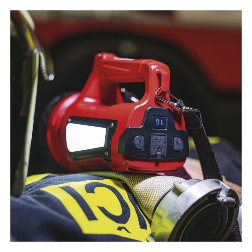 Červená prenosná lampa s displejom ukazuje 96%. Lampa leží na uniforme hasiča s číslom 12. Na pozadí je hasičské auto.