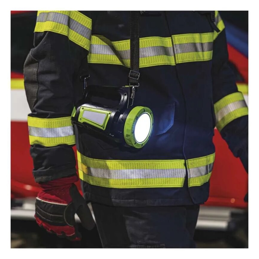 Hasič s baterkou na tele pri zásahe. Baterka svieti. Pozadie tvorí hasičské auto.