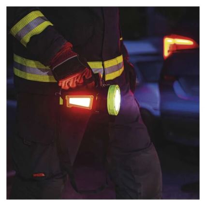 Hasič drží baterku s červeným a bielym svetlom pri dopravnej nehode v noci. Baterka je pripevnená k opasku. Na pozadí sú viditeľné rozmazané svetlá áut.
