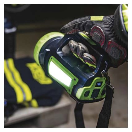 Ruka v ochrannej pracovnej rukavici drží prenosnú baterku s integrovanou LED lampou. Baterka je zeleno-čierna s popruhom. Na pozadí sú ďalšie predmety, pravdepodobne zásahová výbava.