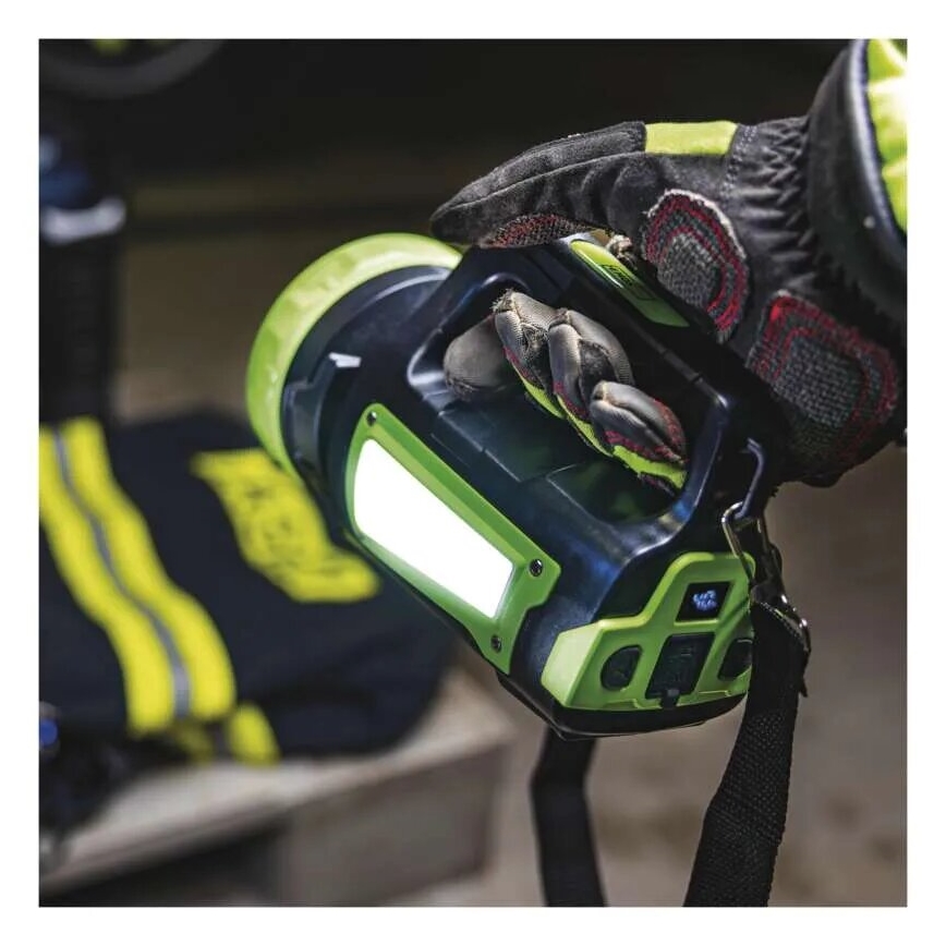 Ruka v ochrannej pracovnej rukavici drží prenosnú baterku s integrovanou LED lampou. Baterka je zeleno-čierna s popruhom. Na pozadí sú ďalšie predmety, pravdepodobne zásahová výbava.