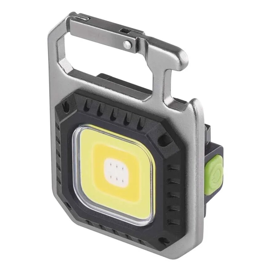 Malá prenosná LED lampa s karabínou svieti. Je štvorcového tvaru, kovového vzhľadu, s čiernymi a sivými prvkami, a má zelené tlačidlo.