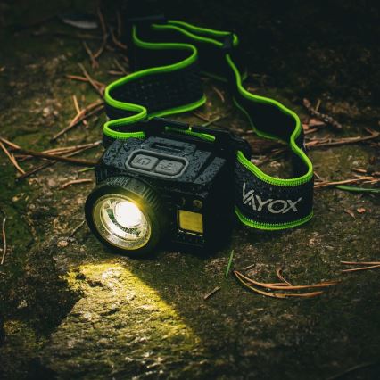 Čelová lampa leží na zemi medzi červenými listami a kmeňom stromu. Lampa je mokrá a svieti červeným svetlom.