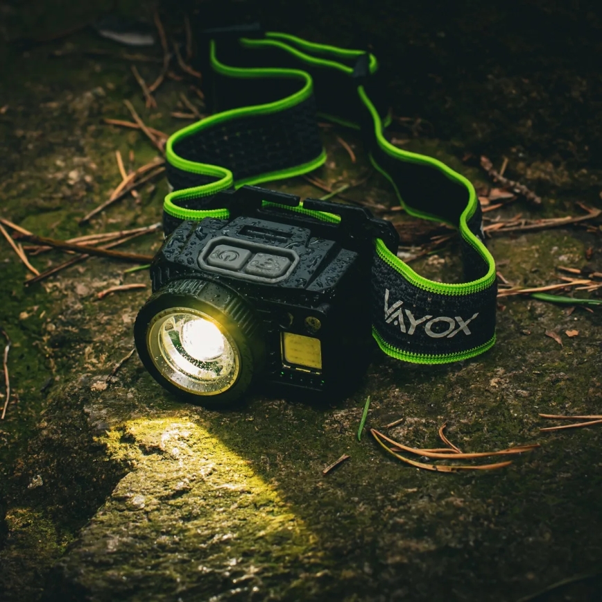 Čelová lampa leží na zemi medzi červenými listami a kmeňom stromu. Lampa je mokrá a svieti červeným svetlom.