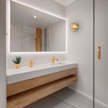 Moderná umývadlová skrinka z dreva a bielej dosky s umývadlom a batériou je umiestnená v kúpeľni. Nad umývadlom je zrkadlo s podsvietením. V kúpeľni je aj sklenená sprchová stena.