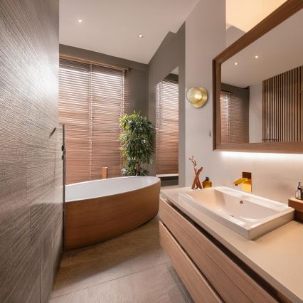 Drevená vaňa stojí v modernej kúpeľni s dreveným nábytkom a žalúziami na oknách. V kúpeľni je aj umývadlo a nástenné svietidlo.