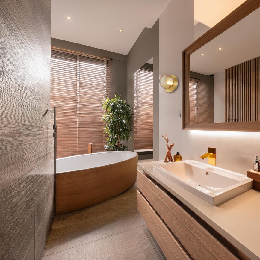 Drevená vaňa stojí v modernej kúpeľni s dreveným nábytkom a žalúziami na oknách. V kúpeľni je aj umývadlo a nástenné svietidlo.