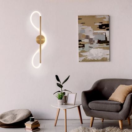 Zlatá nástenná lampa s LED osvetlením v tvare dvoch prepojených slučiek svisle umiestnená na stene. Lampa je súčasťou zariadenia obývacej izby s kreslom, stolíkom a rastlinou.