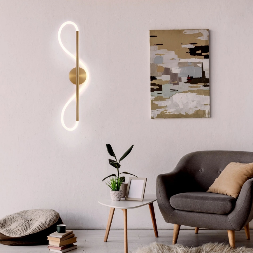 Zlatá nástenná lampa s LED osvetlením v tvare dvoch prepojených slučiek svisle umiestnená na stene. Lampa je súčasťou zariadenia obývacej izby s kreslom, stolíkom a rastlinou.