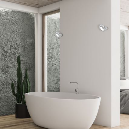 Biela voľne stojaca vaňa sa nachádza v modernej kúpeľni. Dve kovové nástenné svietidlá sú umiestnené na bielej stene. V pozadí je vidieť drsnú textúrovanú sivú stenu a kaktus v kvetináči.