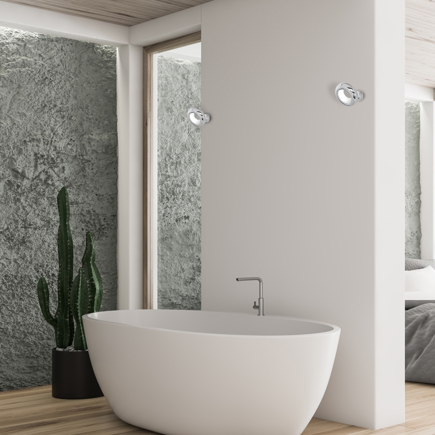 Biela voľne stojaca vaňa sa nachádza v modernej kúpeľni. Dve kovové nástenné svietidlá sú umiestnené na bielej stene. V pozadí je vidieť drsnú textúrovanú sivú stenu a kaktus v kvetináči.
