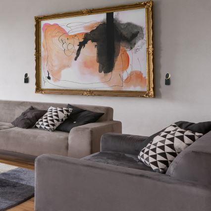 Rohová sedačka s mnohými vankúšmi je umiestnená v obývacej izbe. Na stene sú dve nástenné lampy. Kontext tvorí moderný interiér s drevenou stenou a tmavými závesmi.