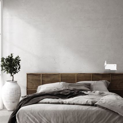 Posteľ s bielou posteľnou bielizňou a vankúšmi je umiestnená v spálni s tehlovou stenou. Na stene je pripevnená biela nástenná lampa. V blízkosti je čierna kovová knižnica.