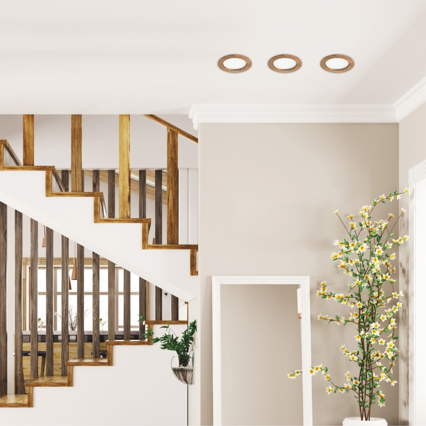 Drevené schodisko vedie nahor v interiéri domu. Na strope sú tri okrúhle svietidlá. V pozadí je vidieť časť jedálne.