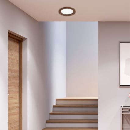 Okrúhle stropné svietidlo s dreveným rámom svieti v interiéri. Nachádza sa v chodbe s drevenými schodmi a dverami. V pozadí je vidieť obraz a nábytok.