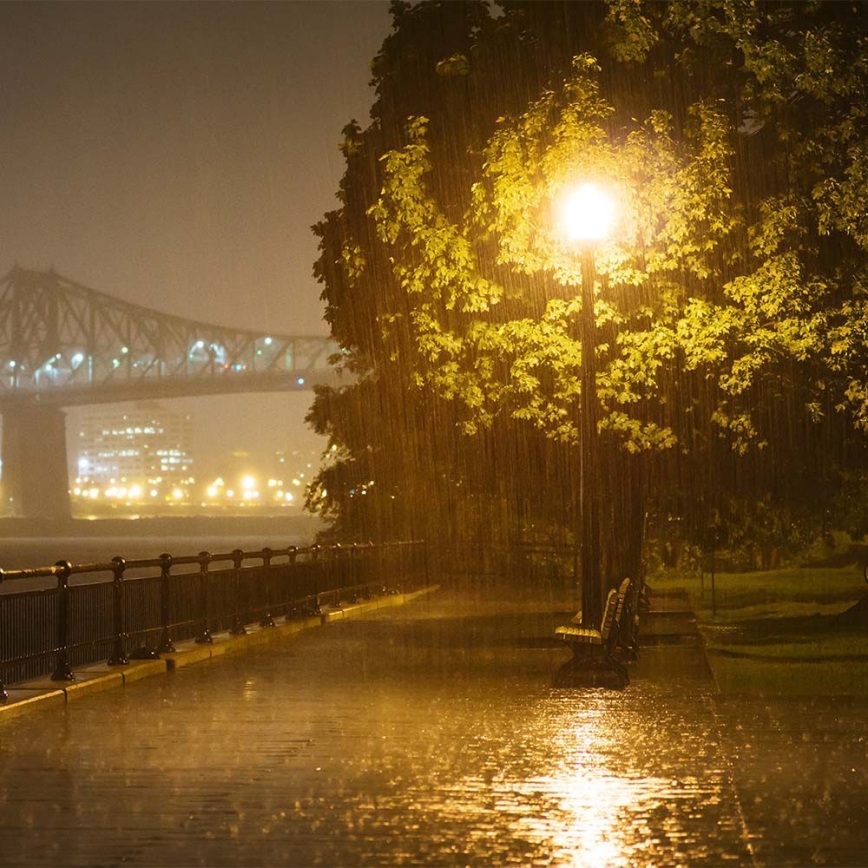 Lampáš osvetľuje dažďom premočený chodník pod stromom pri rieke. V pozadí sa nachádza most a osvetlené mesto.