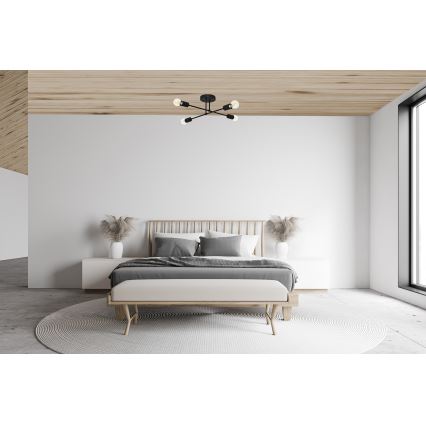 Stropné svietidlo s piatimi ramenami a žiarovkami je umiestnené v spálni. Svietidlo je čierne, svieti v minimalistickej spálni s drevenou posteľou a svetlým nábytkom.