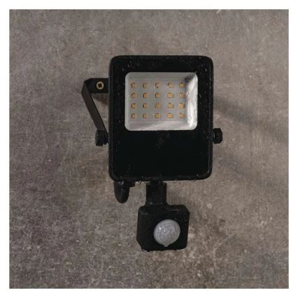 LED reflektor s pohybovým senzorom je pripevnený k stene. Svietidlo obsahuje niekoľko LED diód a je čiernej farby. Senzor je umiestnený pod svietidlom.