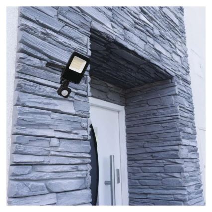 Čierna vonkajšia LED lampa s pohybovým senzorom je pripevnená na sivú kamennú stenu pri vchodových dverách.