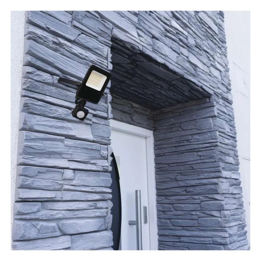 Čierna vonkajšia LED lampa s pohybovým senzorom je pripevnená na sivú kamennú stenu pri vchodových dverách.