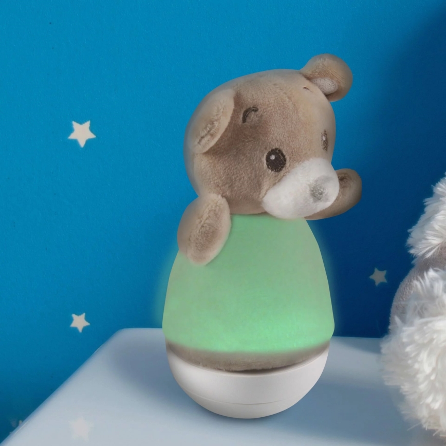 Plyšový medvedík v tvare nočnej lampy svieti jemným zeleným svetlom. Stojí na bielom povrchu pred modrou stenou s malými hviezdičkami. Vedľa neho je časť inej plyšovej hračky.