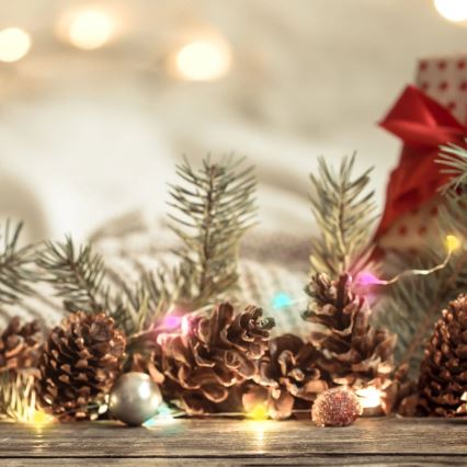 Šišky a ihličie sú rozložené na drevenom povrchu. Sú osvetlené farebnými svetielkami. V pozadí je vlnený materiál a čiastočne viditeľný vianočný darček s červenou mašľou.