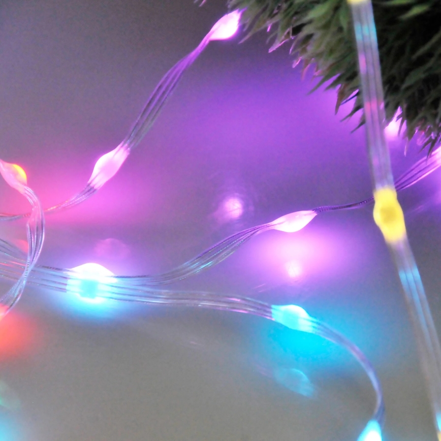 LED svetelný pás svieti viacfarebne. Svetlá sú umiestnené na priehľadnom kábli a vyžarujú fialové, modré a oranžové svetlo. Pás je položený na svetlom povrchu, čiastočne prekrýva umelú vianočnú ratolesť.