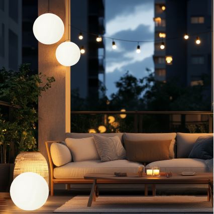 Tri guľové závesné lampy svietia na balkóne. Sediačka s vankúšmi je osvetlená. Vonkajšie osvetlenie vytvára príjemnú atmosféru.