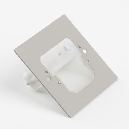 Senzor pohybu je umiestnený na štvorcovej kovovej doske. Doska je svetlo sivej farby a obsahuje otvor pre svetlo. Na doske je umiestnený senzor pohybu s malou šošovkou. Celok leží na bielom podklade.