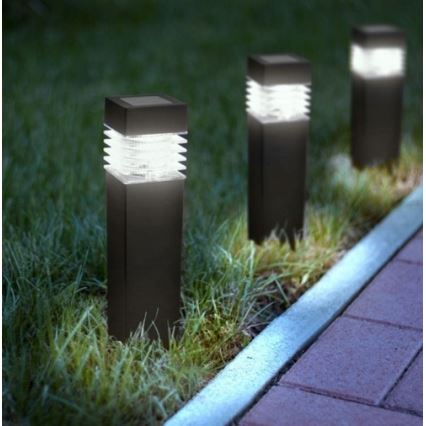 Tri záhradné solárne lampy svietia v tráve popri chodníku. Lampa je vysoká, štvorcová, tmavej farby a svieti teplým svetlom.